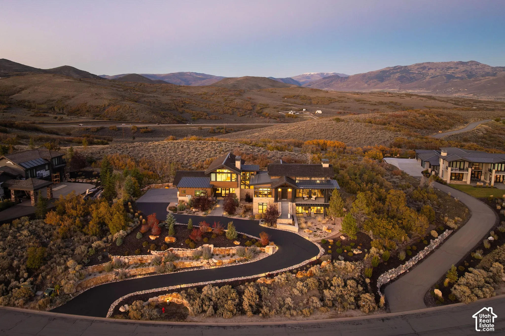 Multi-level architectural residence featuring a metal roof, stone and timber exterior cladding, multi-tier hardscaped retaining walls, paved winding driveway, and mountain scenery