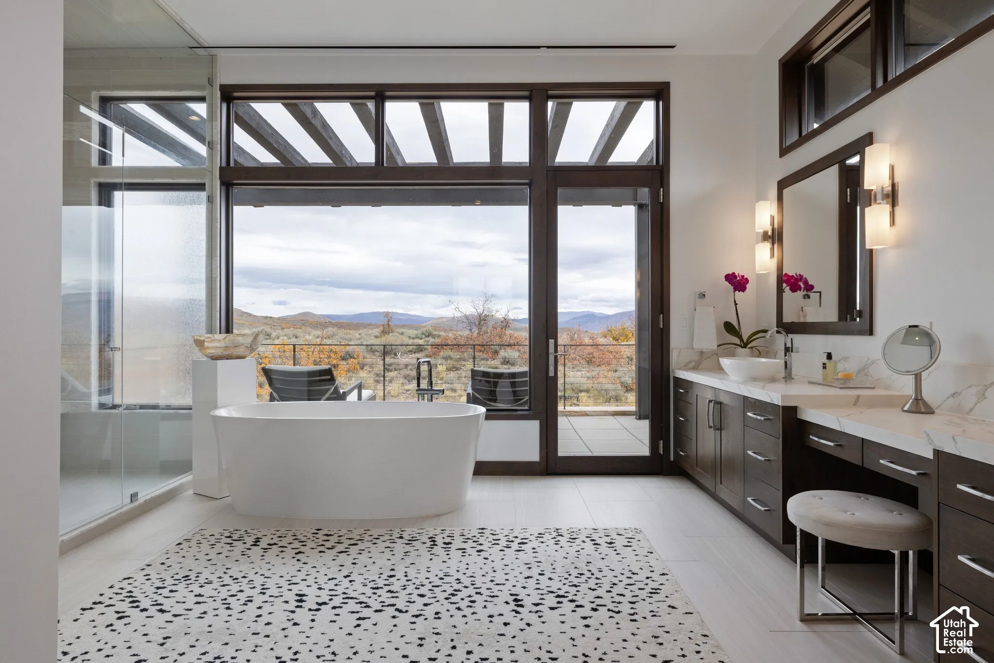 Freestanding soaking tub, floor-to-ceiling windows with mountain scenery, dual-sink vanity with stone-surface countertops, walk-in glass-enclosed shower, and dark-finish cabinetry
