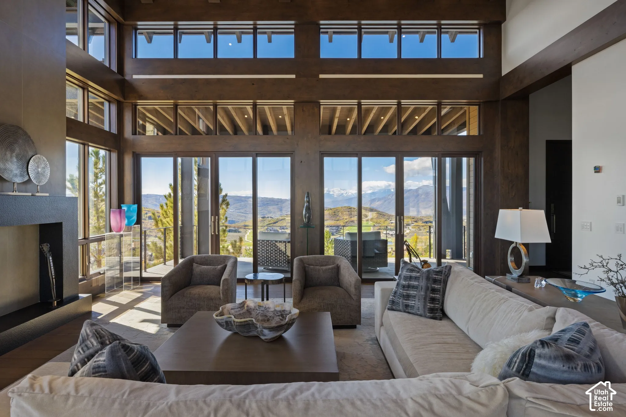 Double-height ceiling with exposed wood beams, expansive floor-to-ceiling windows, mountain scenery, and a minimalist fireplace with a floor-to-ceiling surround