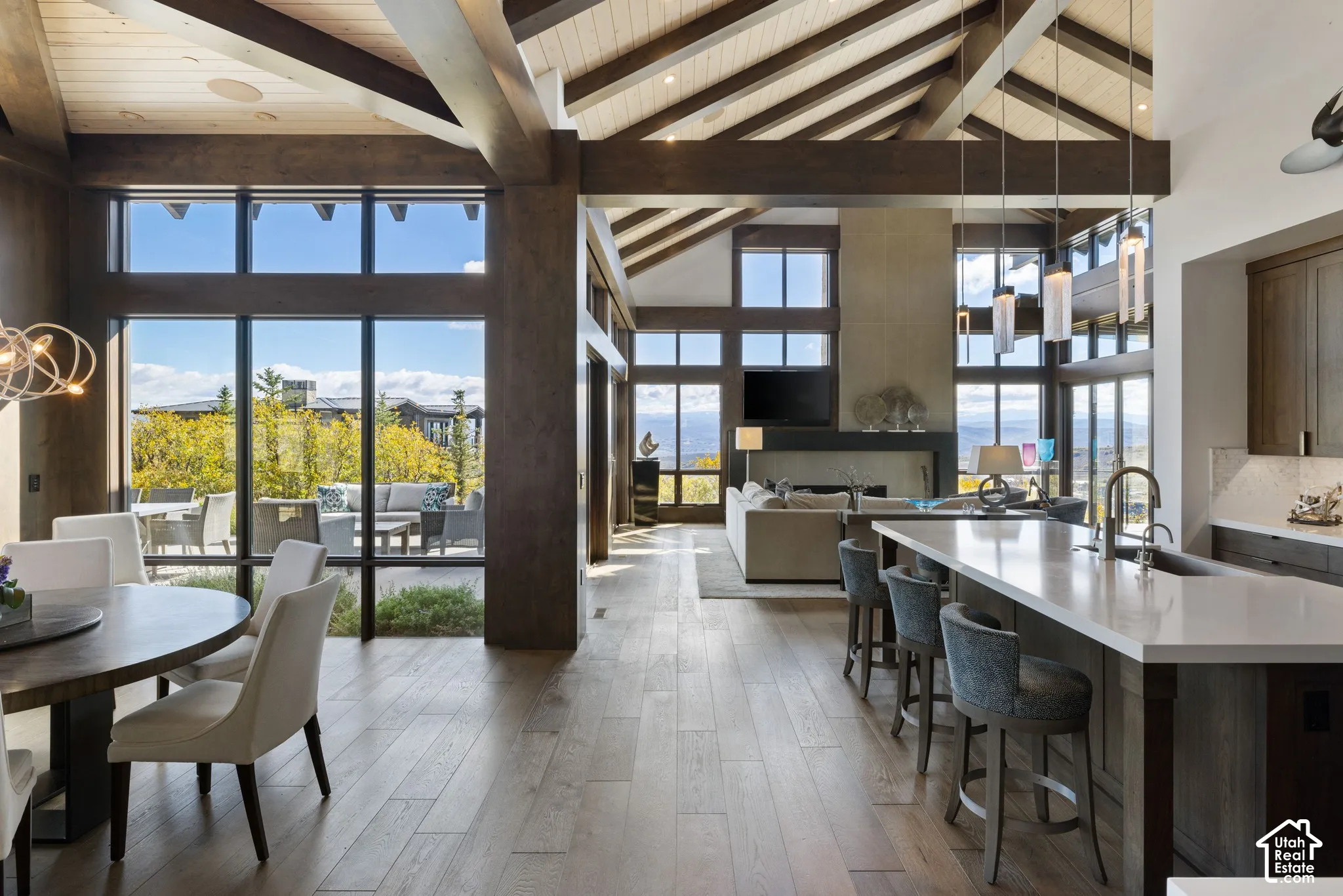 Vaulted ceilings featuring exposed timber beams, expansive floor-to-ceiling windows, wood-finish flooring, a kitchen island with stone-surface countertop, and a floor-to-ceiling fireplace surround