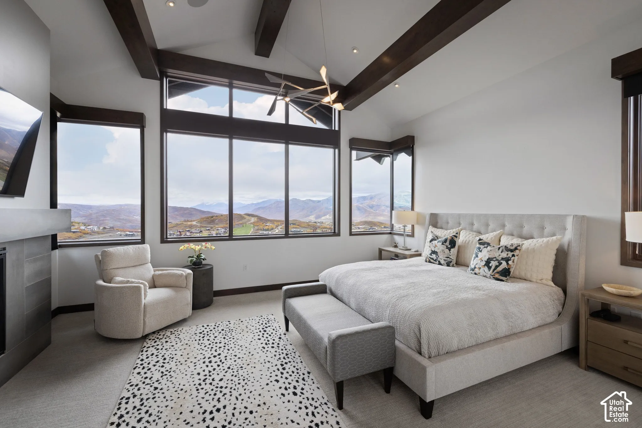 Vaulted ceilings with exposed wood beams, expansive floor-to-ceiling windows, mountain scenery, and a modern metal-surround fireplace