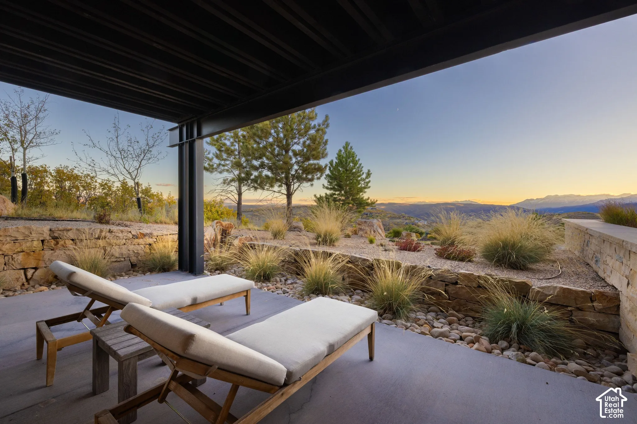 Covered concrete patio featuring structural steel beams, natural stone retaining walls, xeriscaped garden beds, and mountain scenery
