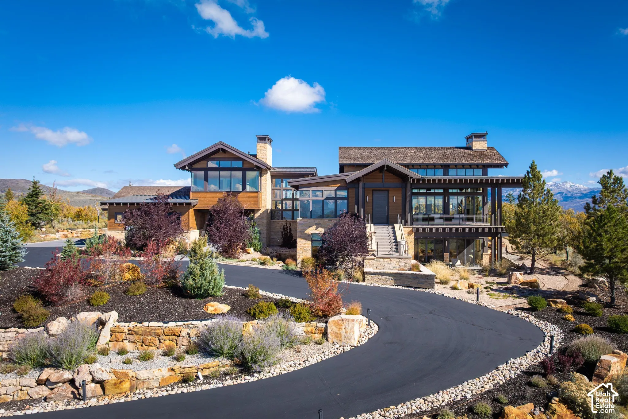 Multi-level exterior featuring stone and wood siding, expansive glass windows, mountain backdrop, and a paved circular driveway