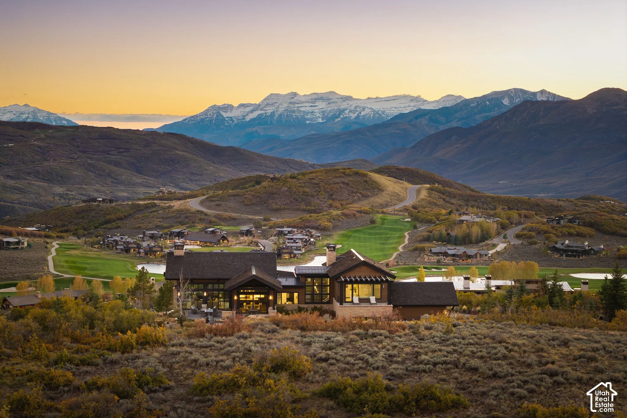 Mountain estate featuring dark wood siding, stone accents, expansive floor-to-ceiling windows, dual stone chimneys, and metal roofing, set against a backdrop of snow-capped peaks