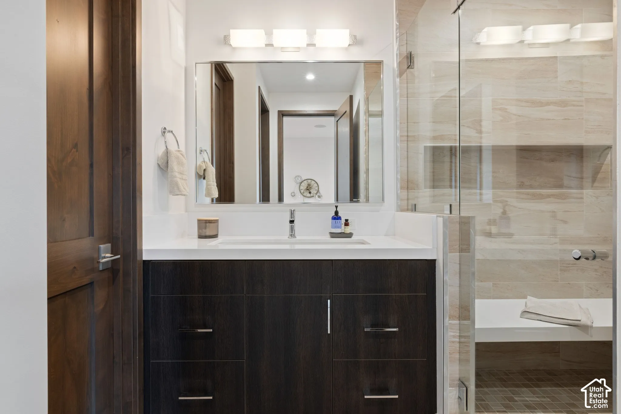 Dark wood-finish vanity with a white solid surface countertop, rectangular under-mount sink, chrome faucet, frameless glass shower enclosure, and built-in shower bench