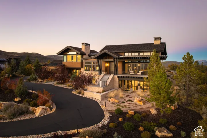 Multi-level mountain modern exterior featuring natural stone and wood siding, steep gabled rooflines, extensive transom windows, elevated balcony, and paved circular driveway
