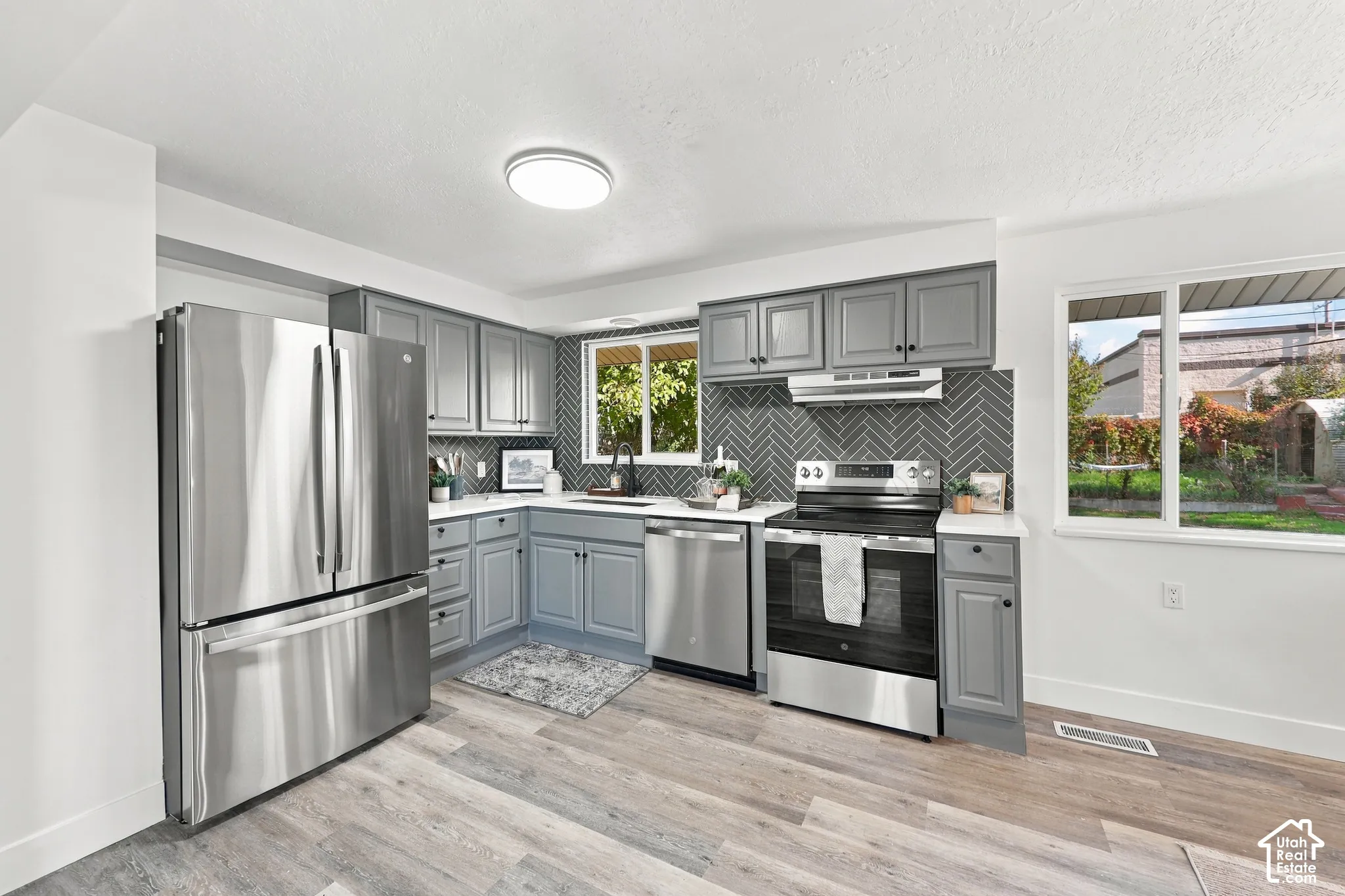 Kitchen featuring grey shaker-style cabinetry, herringbone-pattern tile backsplash, stainless steel appliances, white countertops, and wood-finish flooring