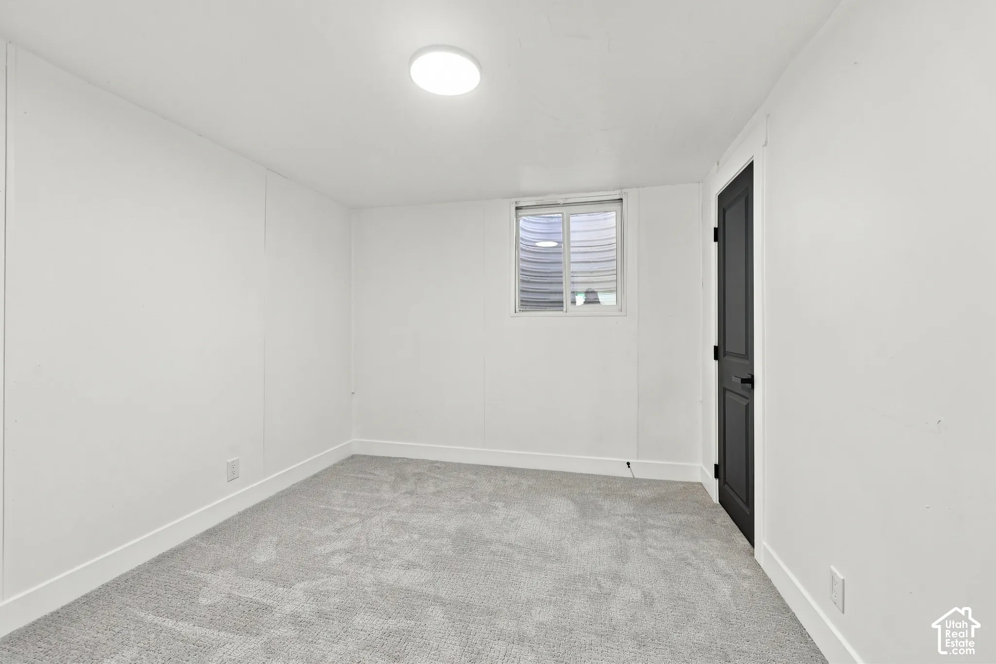 Neutral-toned carpeting, egress window with a window well, and a solid-panel door with matte black hardware