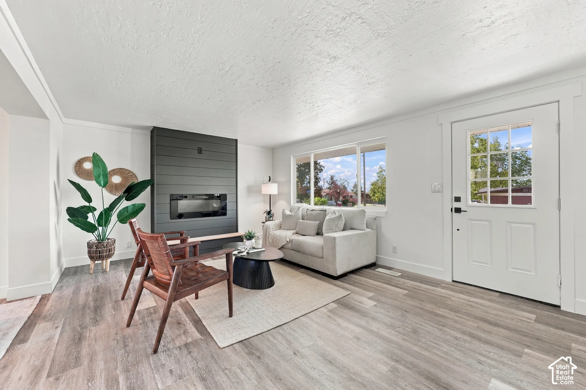 Living area featuring wood-finish flooring, a wall-mounted electric fireplace with a dark shiplap surround, large window with natural light, and a white exterior-style entry door with a multi-pane window insert