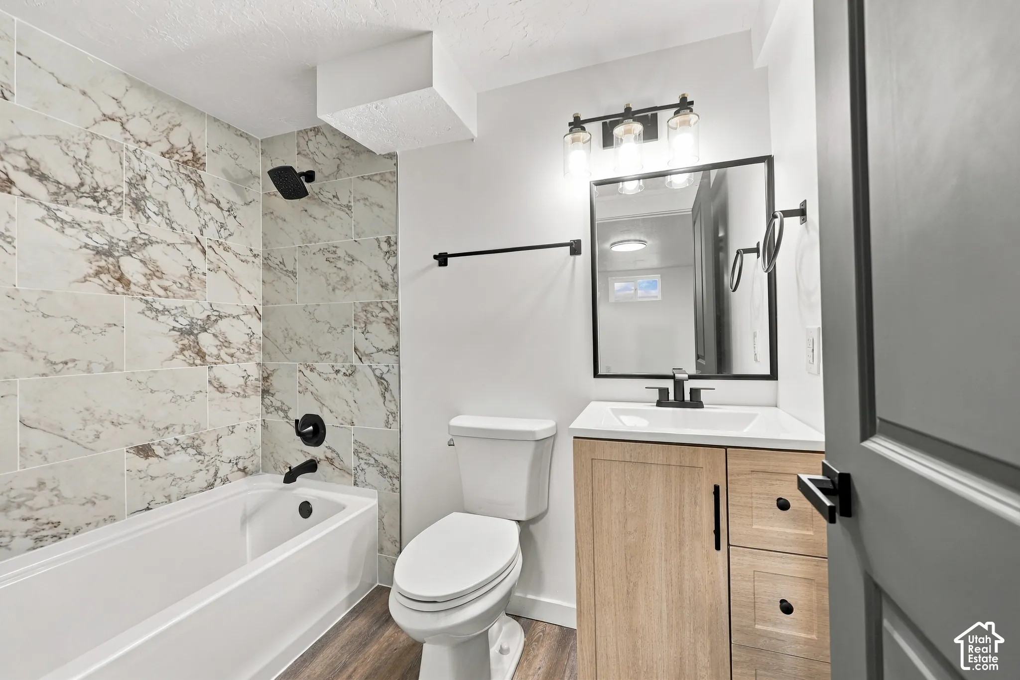 Bathroom featuring a tub-shower combination with floor-to-ceiling patterned wall tile, wood-finish vanity with a white countertop, matte black fixtures, and a framed vanity mirror with three-light vanity lighting