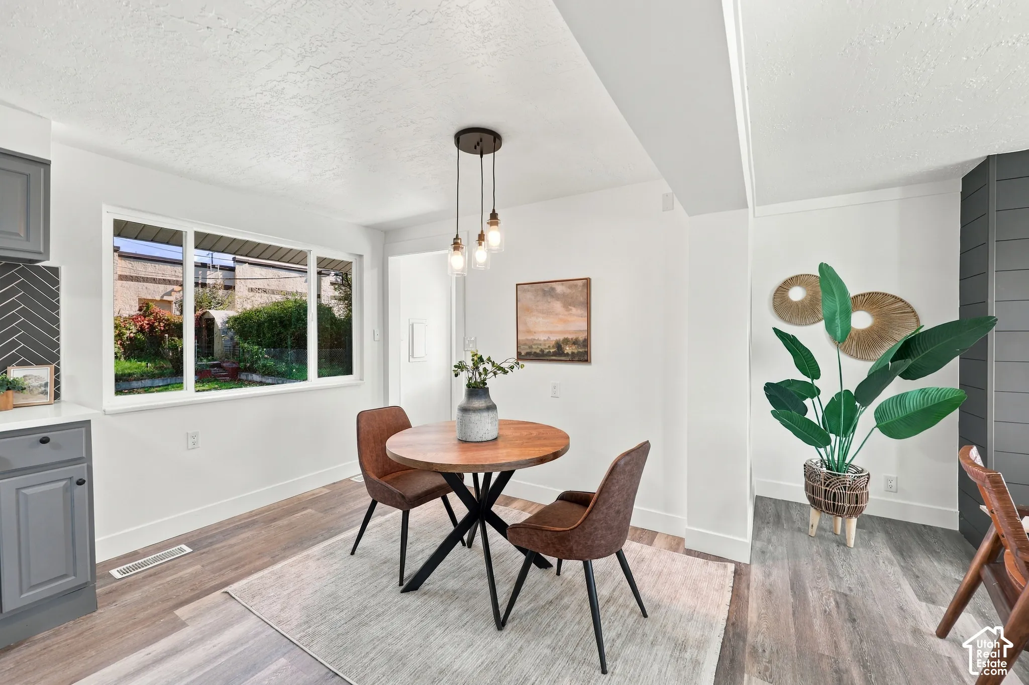 Neutral-toned kitchen area featuring wood-finish flooring, charcoal-toned cabinetry, a herringbone-patterned tile backsplash, and a multi-light pendant fixture