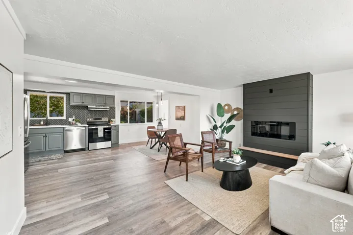 Open-concept interior featuring wood-finish flooring, a kitchen with grey cabinetry and herringbone-patterned backsplash, and a floor-to-ceiling accent wall with a built-in fireplace
