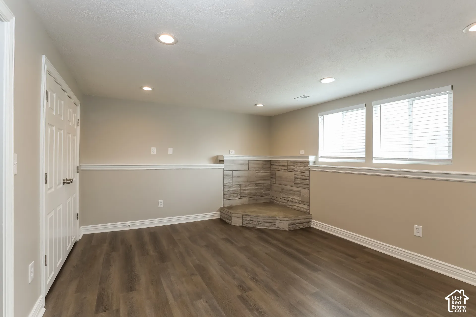 Interior space featuring wood-finish flooring, recessed lighting, chair rail molding, two multi-pane windows with horizontal blinds, and a raised corner hearth with stone-finish surround