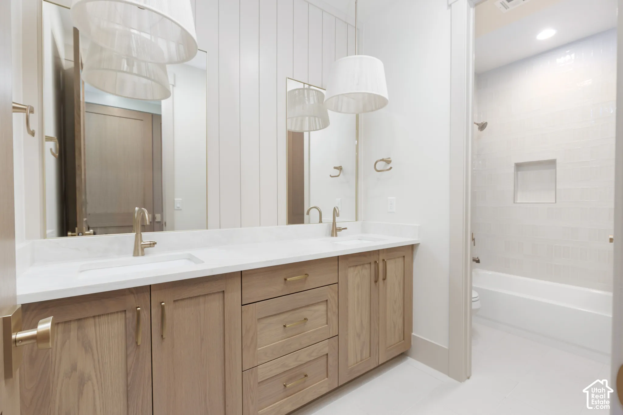 Double vanity featuring light wood-finish cabinetry, white stone-composite countertops, integrated sinks with metallic fixtures, vertical shiplap wall paneling, and pendant lighting