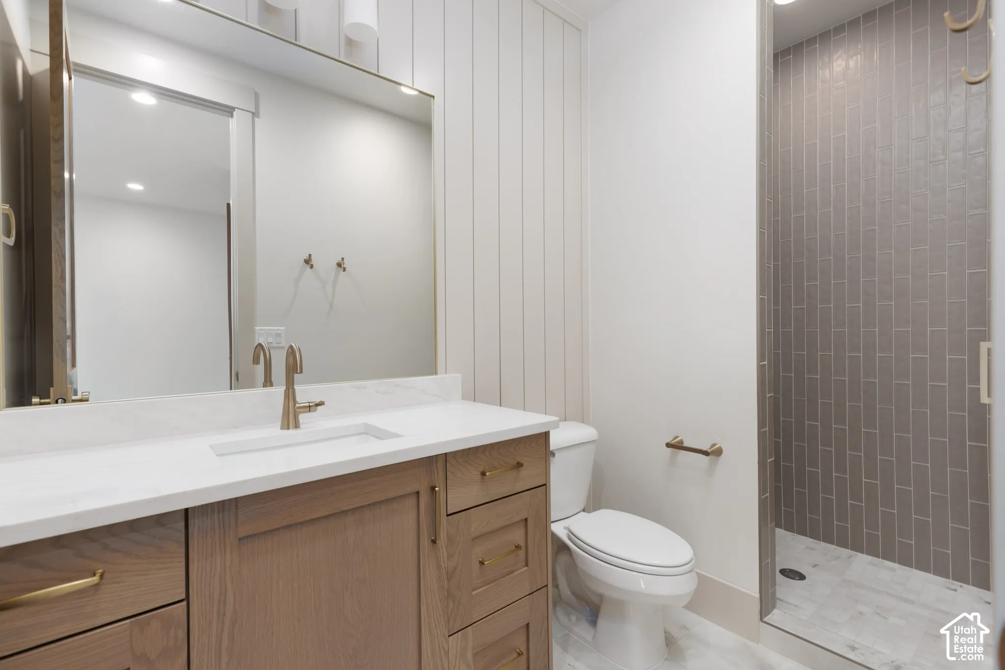 Wood-finish cabinetry with brass hardware, white stone-surface countertop, full-height vertical wall paneling, and a walk-in shower with vertical tile surround