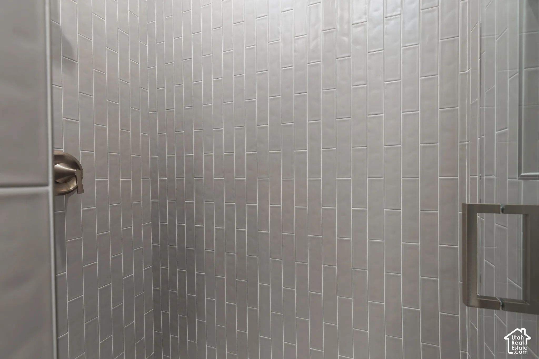 Shower enclosure featuring vertical light-gray subway tile, satin-finish metal plumbing fixtures, and a glass door with a square metal handle