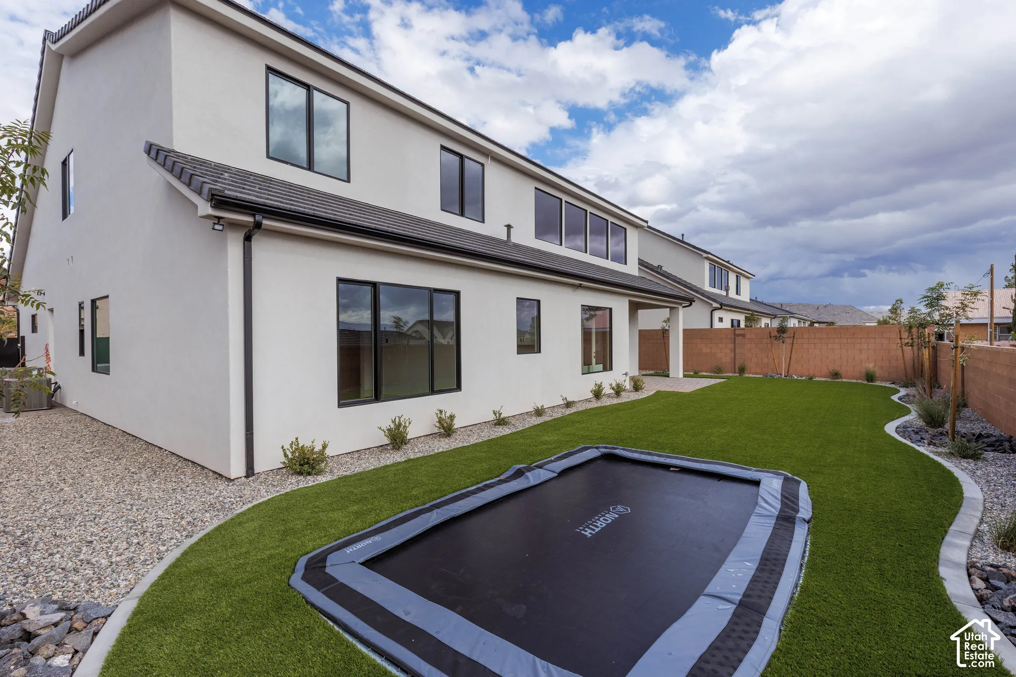 Stucco exterior featuring dual-story height, dark-framed windows, synthetic turf lawn, stone-border hardscaping, and a block-wall perimeter