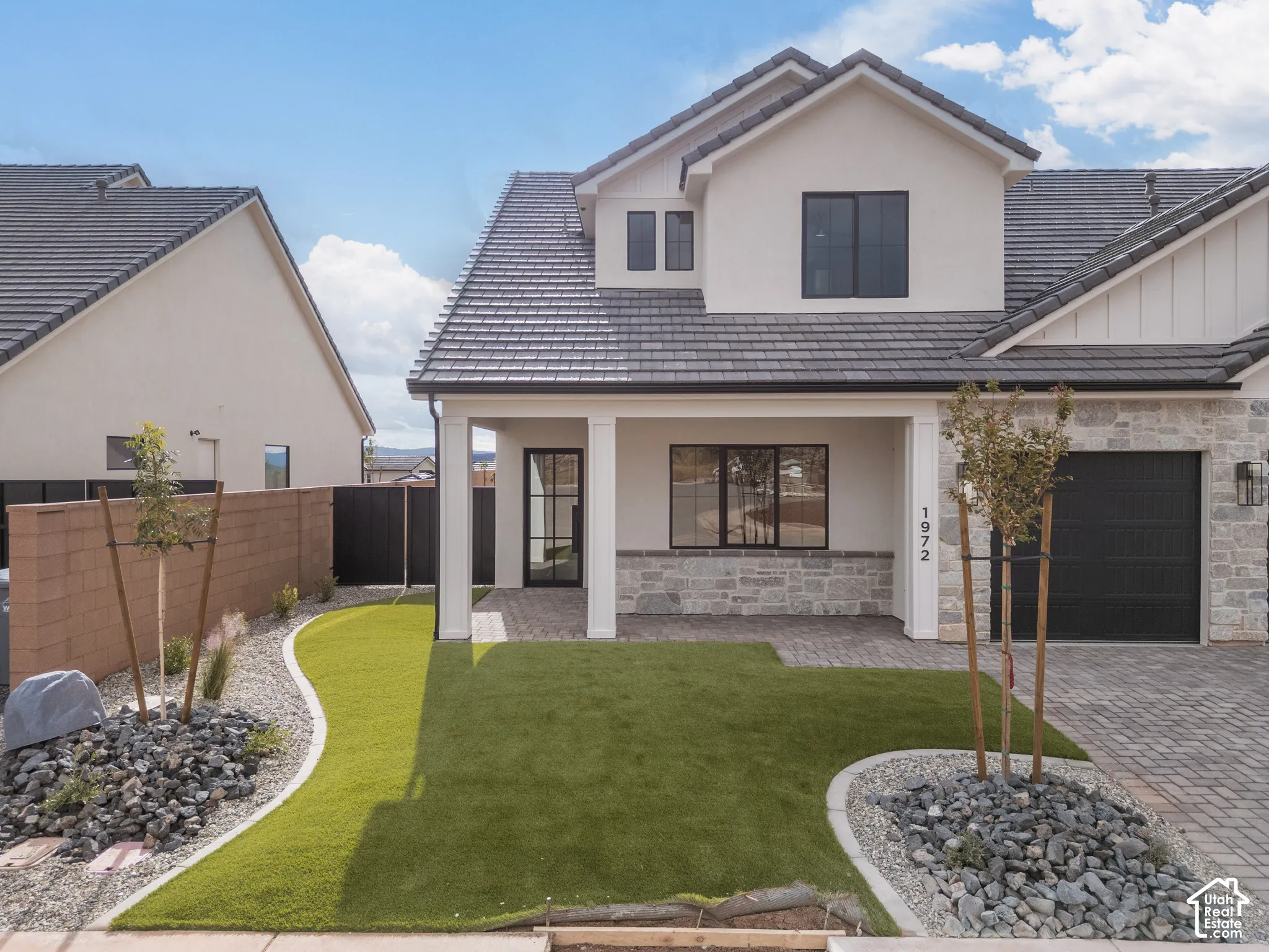 Two-story exterior featuring light-toned stucco and stone facade, dark-toned tile roof, paver driveway, covered front entryway, and landscaped front yard with synthetic turf