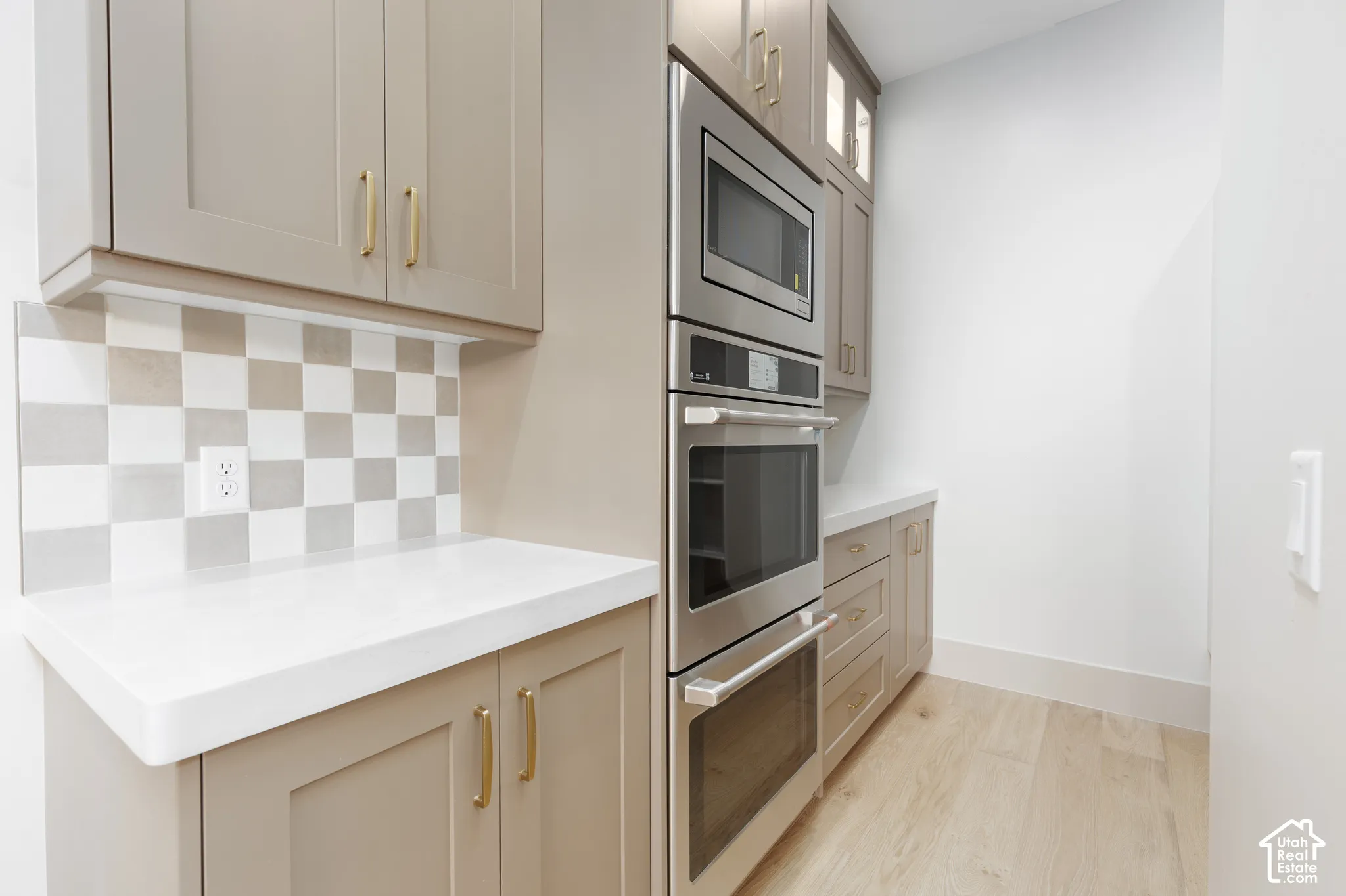 Shaker-style cabinetry with metallic pulls, built-in stainless steel microwave and double oven, light-toned countertop, checkered tile backsplash, and light wood-finish flooring