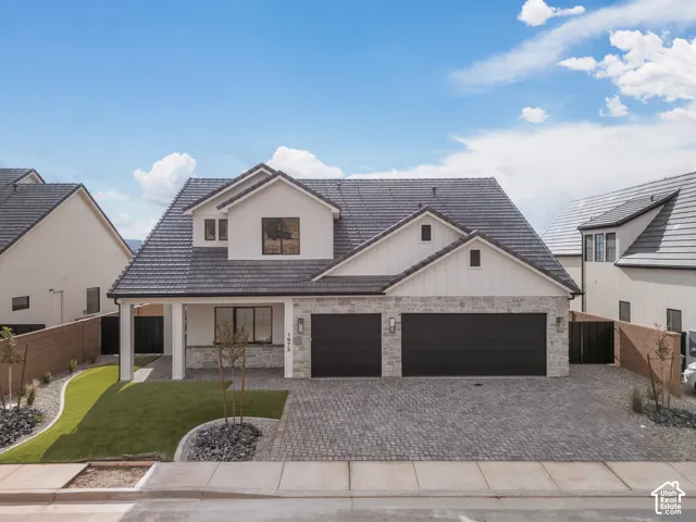 Multi-gabled exterior featuring light stucco, stone veneer, dark concrete tile roofing, a three-car garage, and a cobblestone-patterned paver driveway