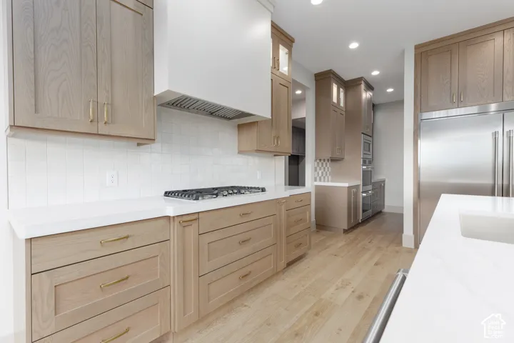 Kitchen featuring light wood-finish cabinetry, solid-surface countertops, a white tile backsplash, stainless steel appliances, and wood-finish flooring