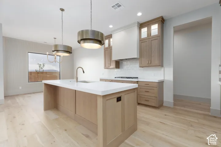 Kitchen featuring light-tone wood cabinetry, a central island with a solid surface countertop, wood-finish flooring, two large cylindrical pendant lights, and a custom range hood