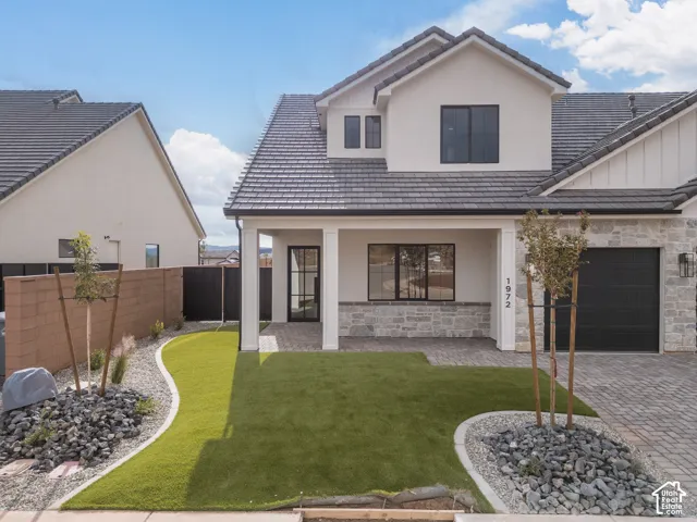 Two-story exterior featuring light-toned stucco and stone facade, dark-toned tile roof, paver driveway, covered front entryway, and landscaped front yard with synthetic turf