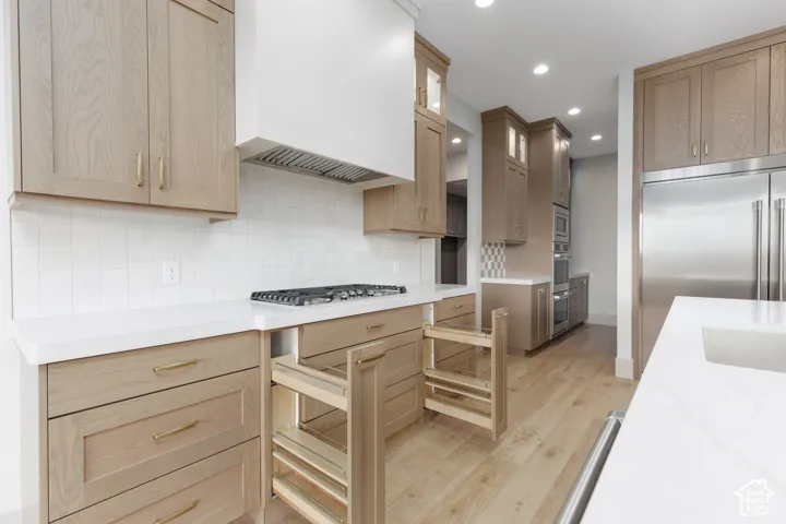 Kitchen featuring light-tone wood cabinetry, solid-surface countertops, built-in stainless steel appliances, a gas cooktop with a custom range hood, and wood-finish flooring