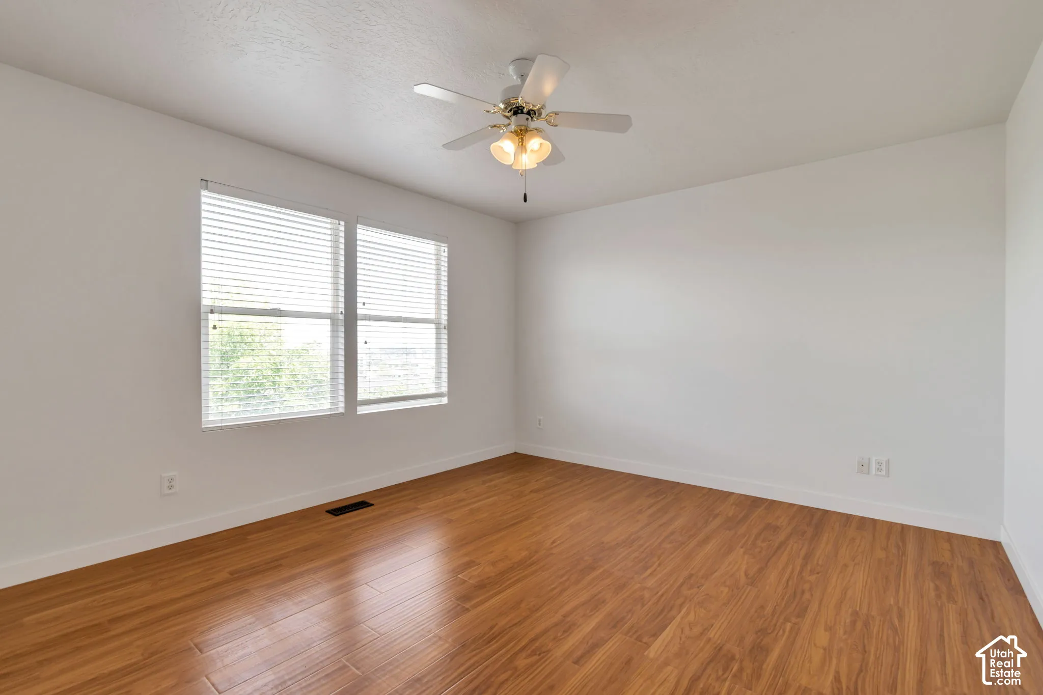 Wood-finish flooring, white walls, dual pane windows with horizontal blinds, and a central ceiling fan with integrated lighting