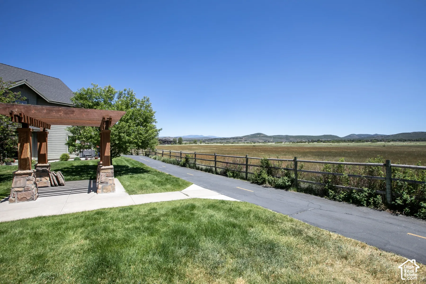Outdoor patio space featuring a wooden pergola with stone column bases, concrete paving, manicured lawn, and post-and-rail fencing with mountain backdrop
