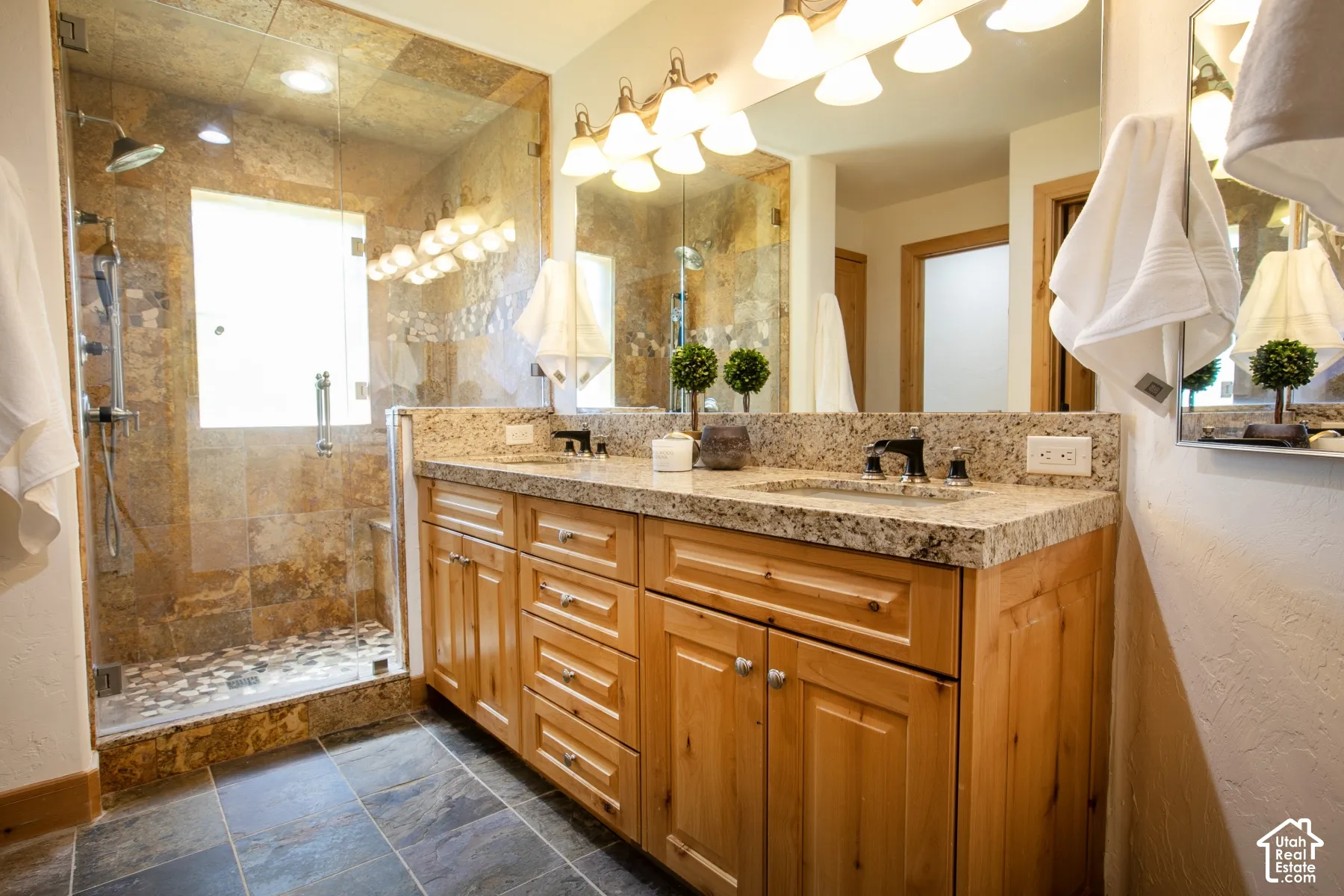 Bathroom featuring a dual-sink vanity with light-toned wood cabinetry, stone-finish countertop, glass-enclosed walk-in shower with stone tile surround, and large wall-mounted vanity mirror