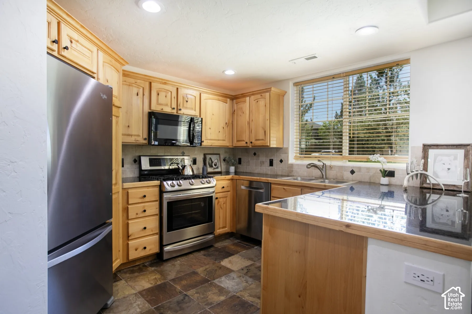 Kitchen featuring natural wood cabinetry, dark stone countertops, stainless steel appliances, stone tile flooring, and a double-basin sink