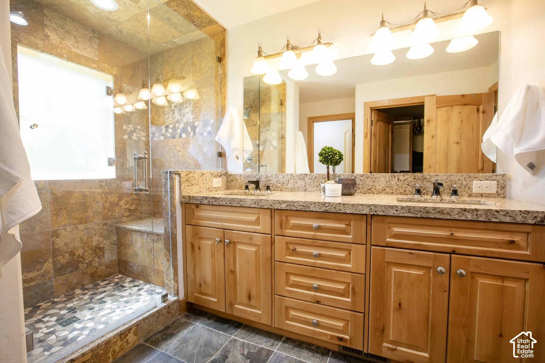 Dual-sink vanity with stone countertop, light wood cabinetry, stone-tiled walk-in shower with integrated bench, and wall-to-wall vanity mirror