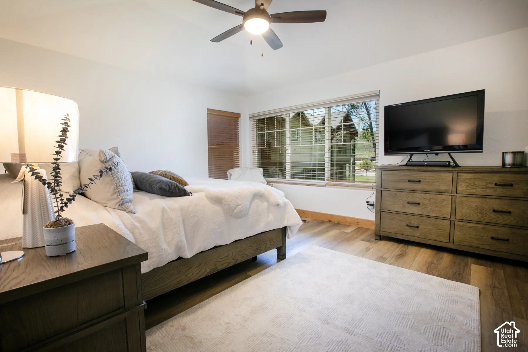 Vaulted ceiling with integrated multi-blade ceiling fan, wood-finish flooring, and large window with slatted blinds