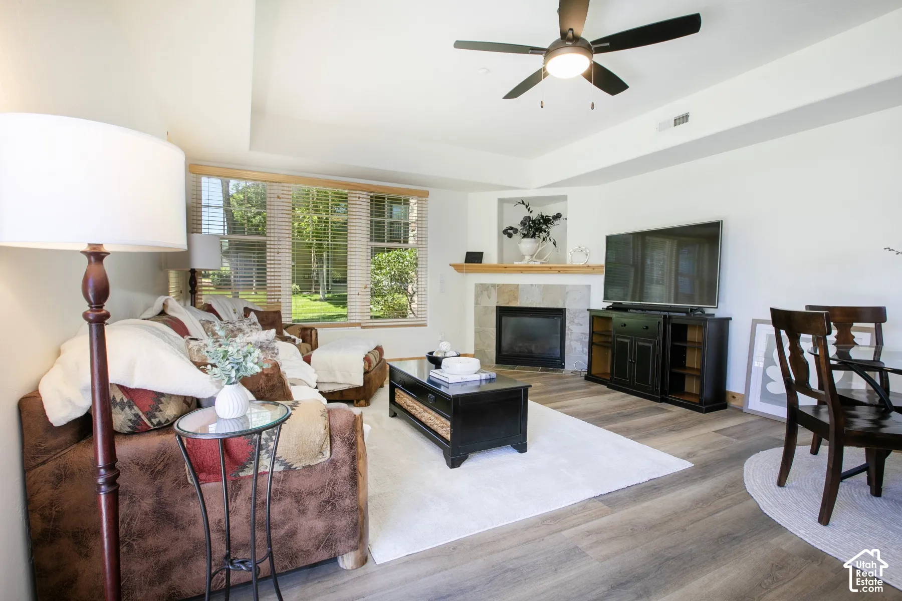 Living area featuring wood-finish flooring, a tray ceiling with a central fan, multi-pane windows, and a stone-surround fireplace with a wooden mantel