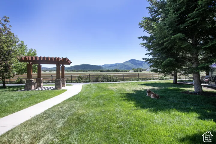Backyard featuring a wooden pergola with stone column bases, a concrete walkway, mature evergreen trees, and perimeter wooden fencing against a mountain backdrop