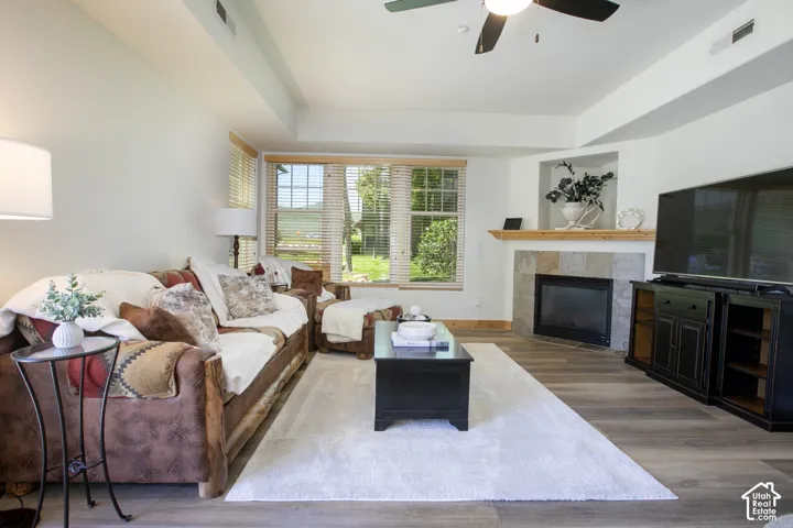 Living room featuring wood-finish flooring, tray ceilings, multi-panel windows, and a stone-surround fireplace with a timber mantel