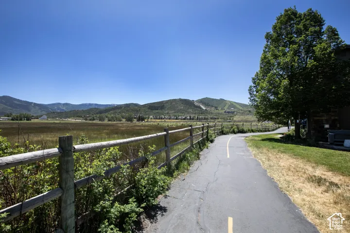 Paved recreation path bordered by a rustic timber-rail fence, open meadow, and mountain backdrop