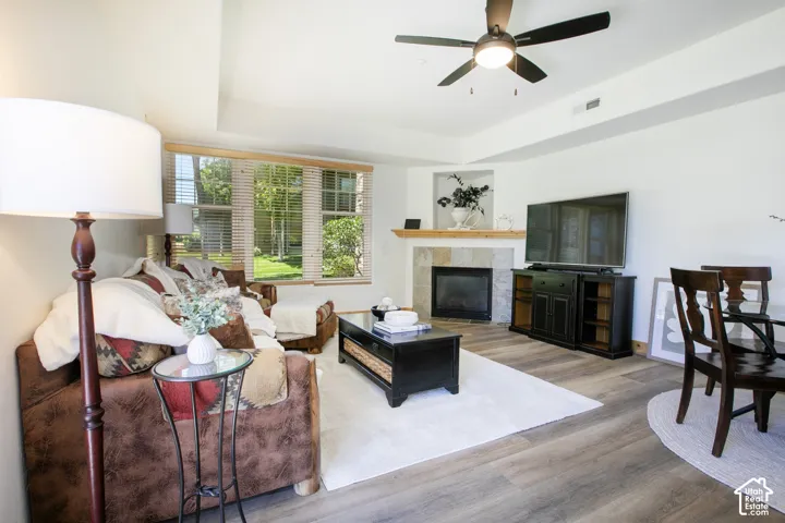 Living area featuring wood-finish flooring, a tray ceiling with a central fan, multi-pane windows, and a stone-surround fireplace with a wooden mantel