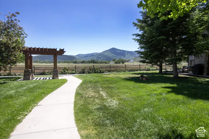 Backyard featuring a grass lawn, stone-pillared wood pergola, concrete pathway, perimeter split-rail fencing, and mature evergreen trees against a mountain backdrop