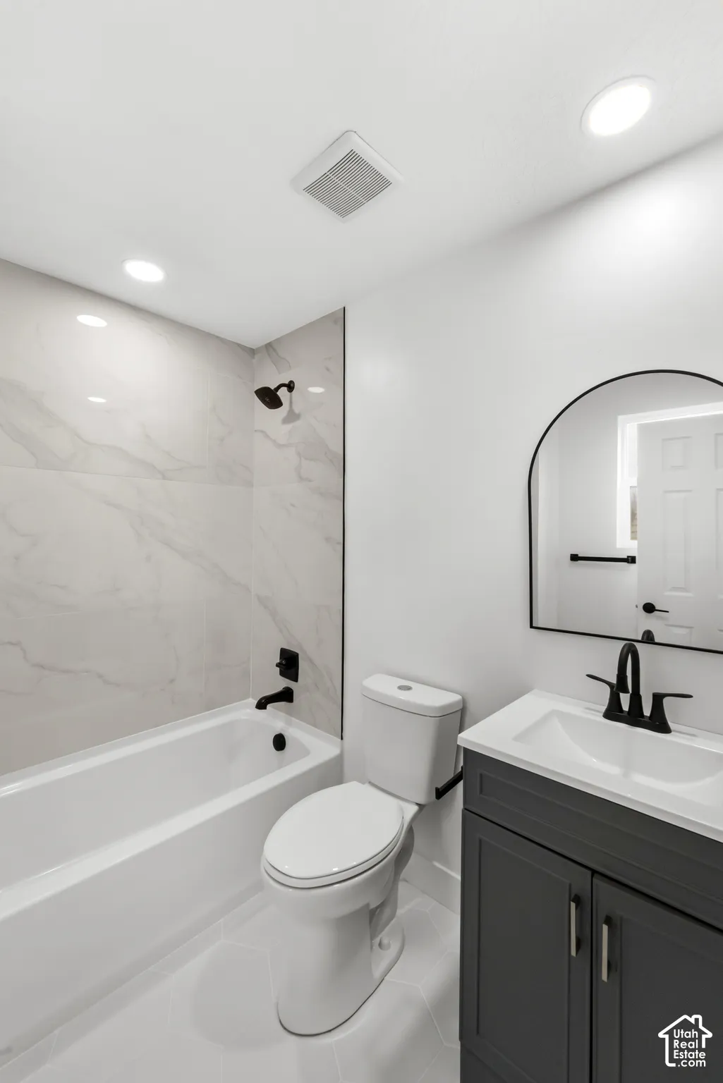 Bathroom featuring a charcoal-finish vanity with a white countertop, arched-frame mirror, matte black fixtures, and a shower-tub combination with light-toned, large-format tile surround