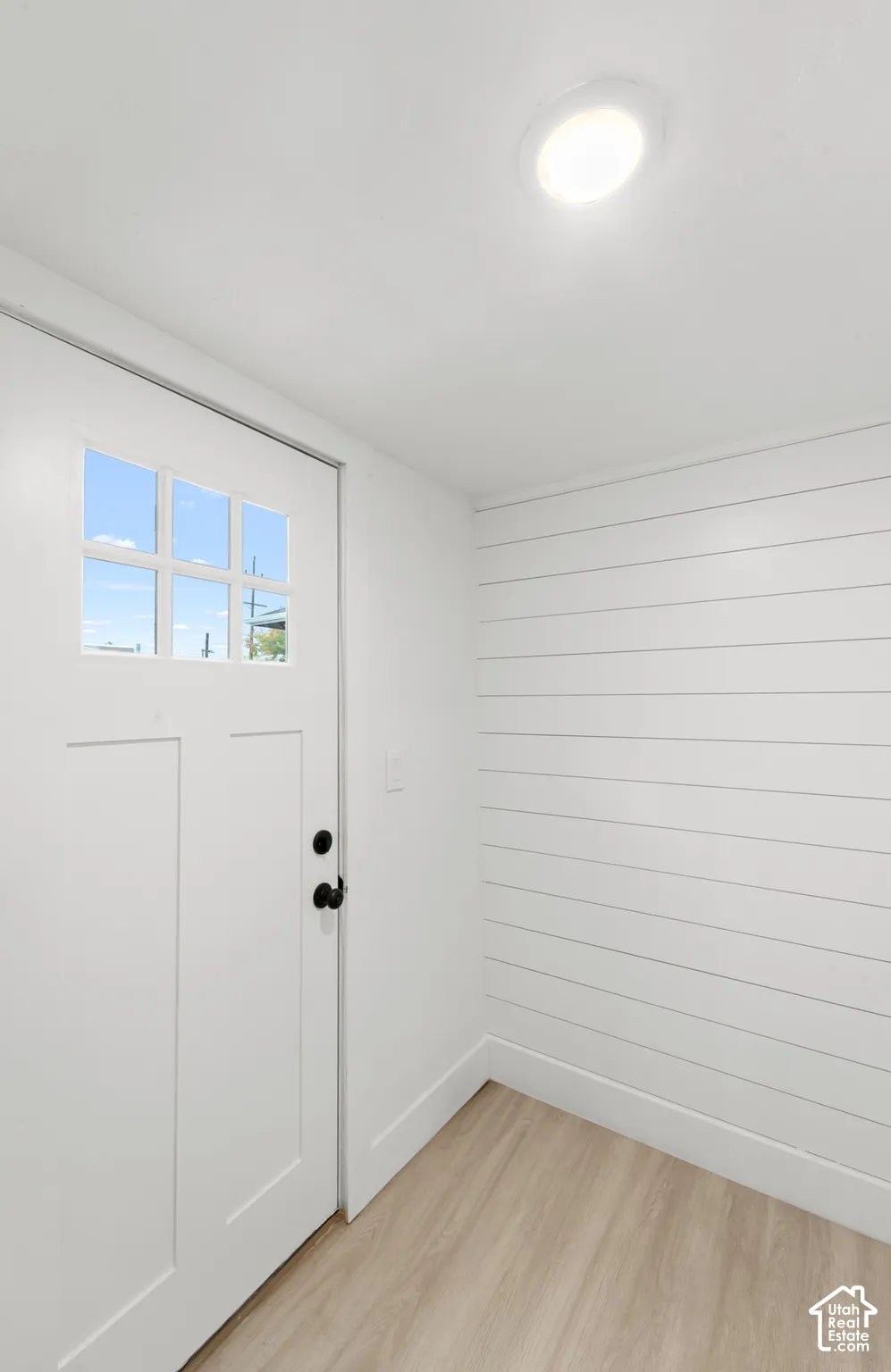 Crisp white shiplap wall paneling, light-tone wood-finish flooring, a recessed ceiling light, and a white six-pane glass insert door with matte black hardware