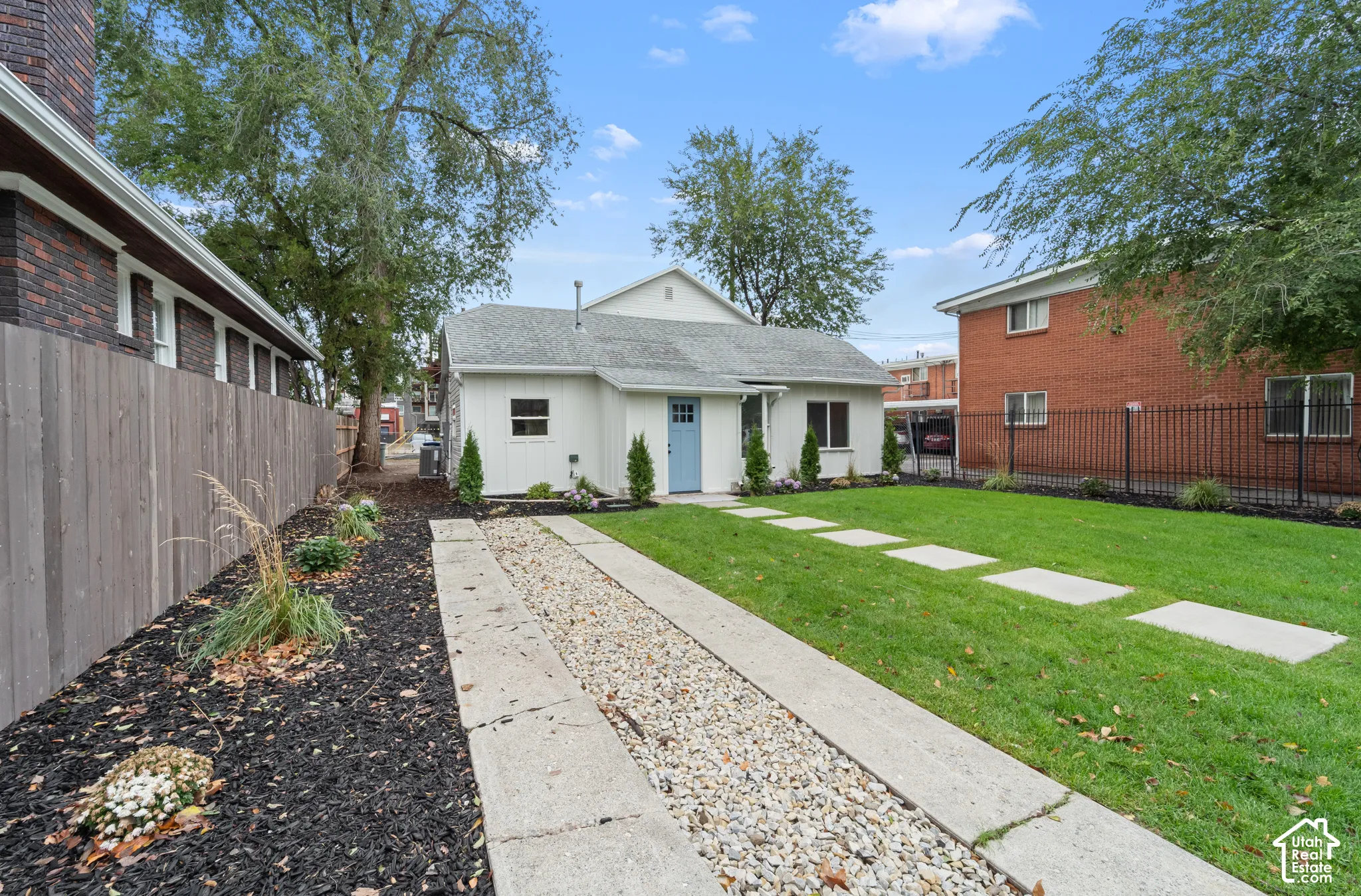 Exterior backyard space featuring a manicured lawn, concrete paver pathway, decorative gravel strip, vertical wood-panel fencing, and mature trees