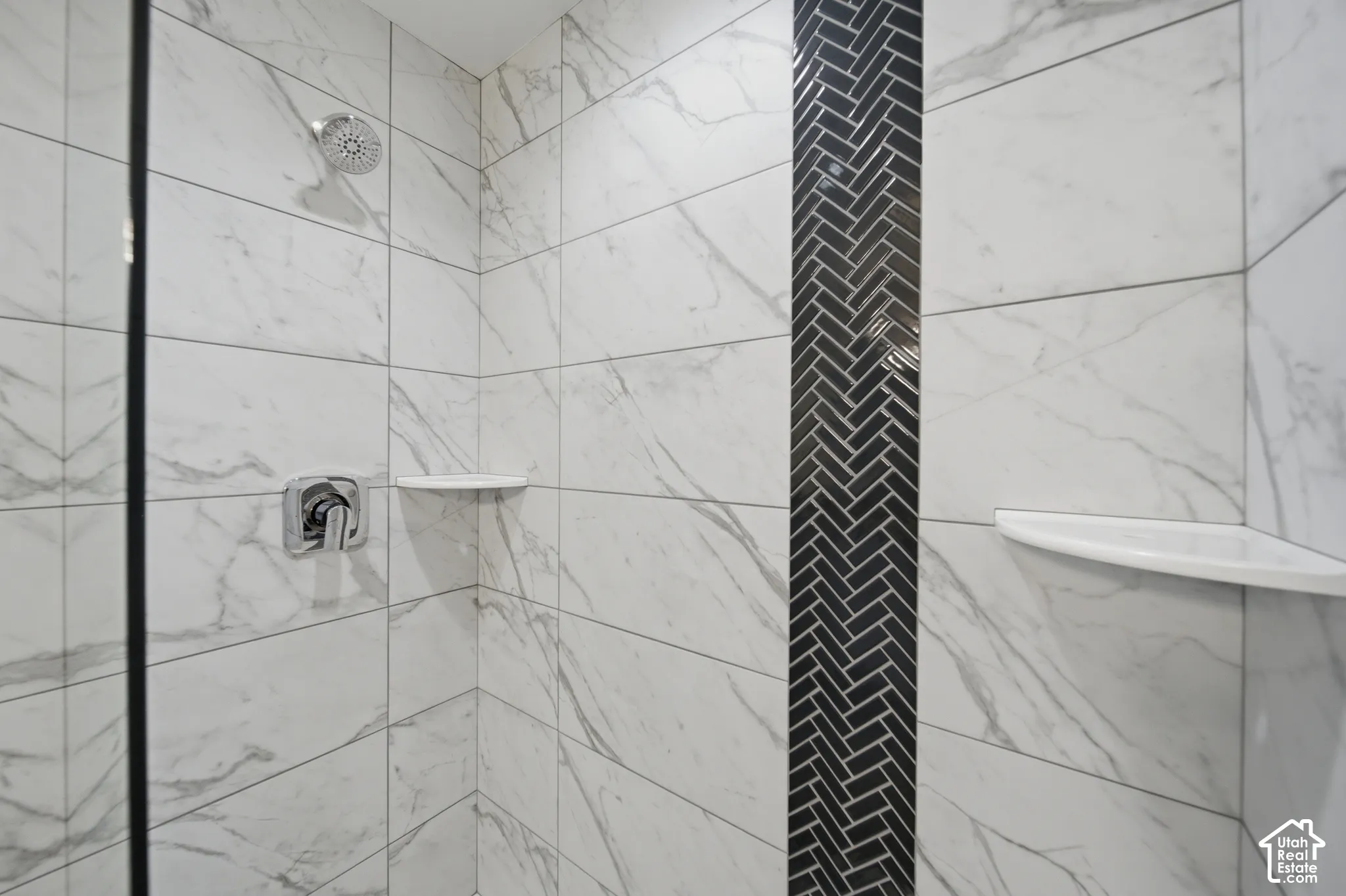 Shower with large format white marble-look tile, a black herringbone accent strip, chrome fixtures, and integrated corner shelving