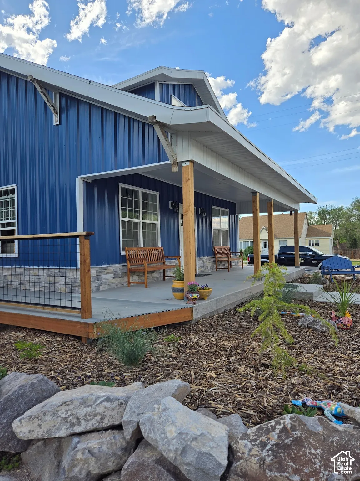Exterior featuring blue vertical metal siding, covered porch with wood support posts, stone veneer skirting, and wire-mesh railing