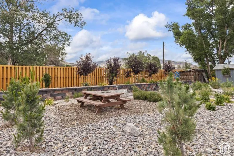 Landscaped backyard featuring stone masonry retaining walls, a wood-slat perimeter fence, rock ground cover, and mature pine trees set against a mountain backdrop