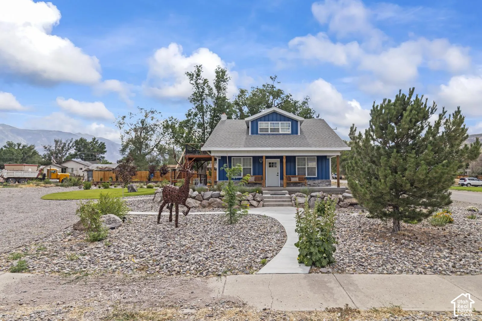 Blue-sided exterior featuring stone-veneer accents, a covered front porch with timber support beams, and a concrete walkway leading to mountain-backed surroundings