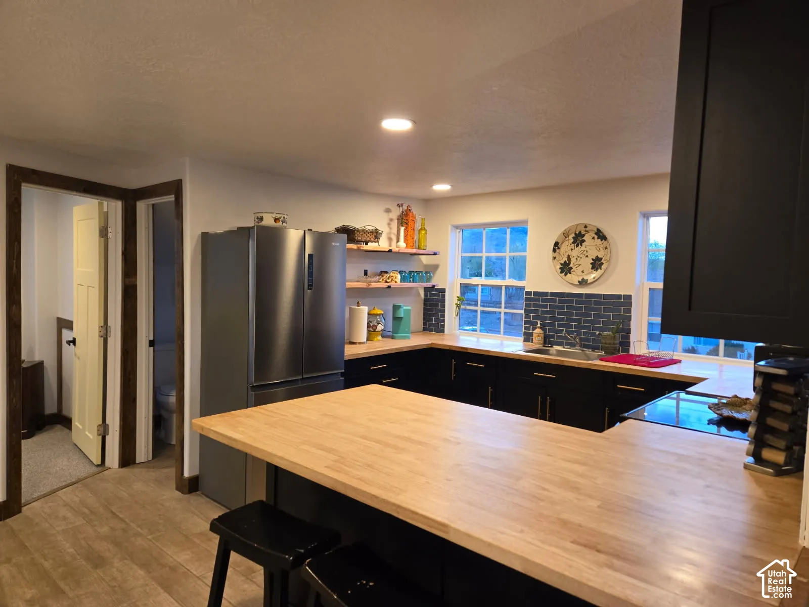Kitchen featuring wood-finish countertops, dark cabinetry, blue tile backsplash, stainless steel refrigerator, and open wood shelving