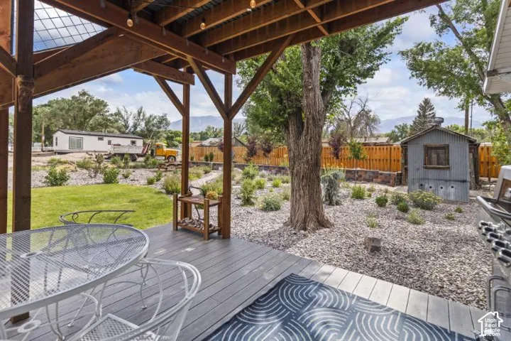 Covered outdoor patio featuring exposed timber framing, composite deck flooring, and mountain backdrop, complemented by a landscaped backyard with a stone-accented perimeter wall and detached metal utility structure