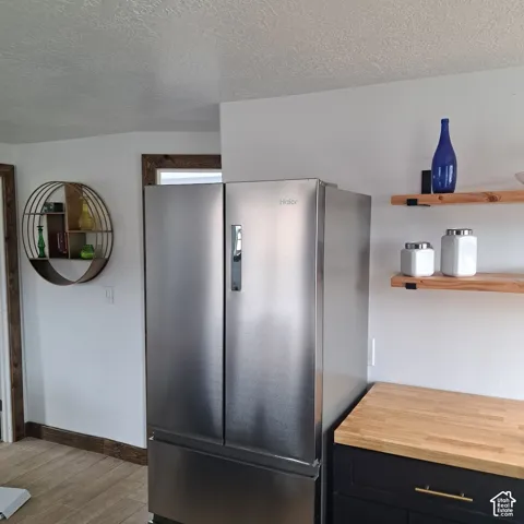 Stainless steel French door refrigerator, wood-finish countertop, dark cabinetry with gold-tone hardware, and natural wood floating wall shelving